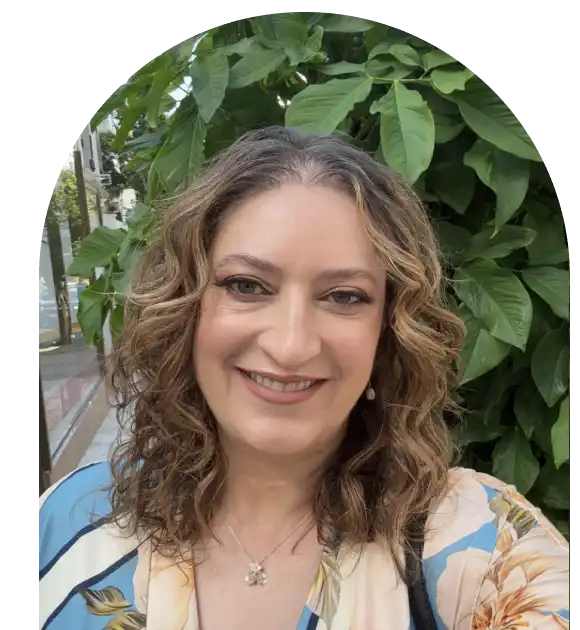 Smiling woman with curly hair in front of leafy green plants.