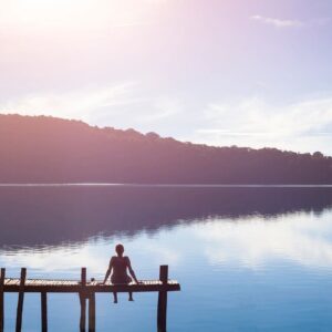 Person sitting on a dock overlooking a calm lake at sunrise.